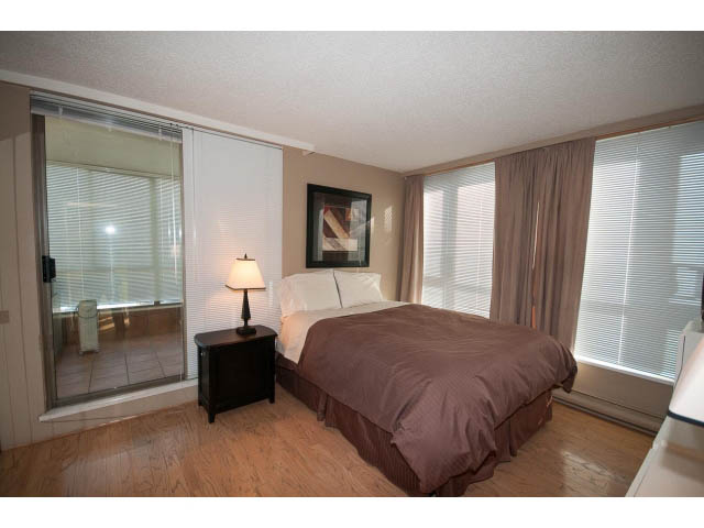 Furnished bedroom with hardwood floors and large windows, featuring a cozy bed and a nightstand with a lamp.