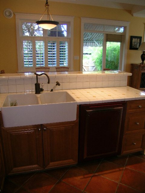 Bright kitchen with white tile countertops and a double sink, featuring a window with shutters and a hanging light fixture.