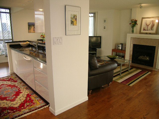 Bright kitchen with black countertops and hardwood floors, leading into a cozy living area with a fireplace and city view.