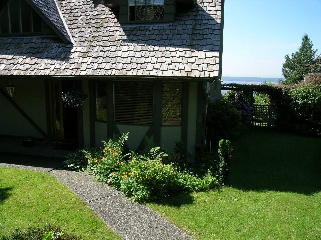 Bright living room with wooden beams, stone fireplace, and lush garden. - 1332 Camridge Place, West Vancouver