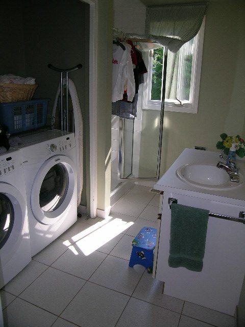 Bright laundry room with white appliances and tiled floor, adjacent to a small bathroom with a sink and window. - 1332 Camridge Place, West Vancouver