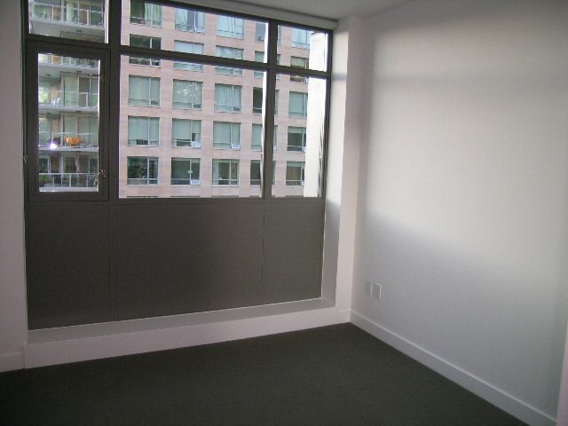 Bright living room with large windows offering a city view, clean white walls, and dark carpet. - 602 - 5782 Berton Avenue, Vancouver West Side