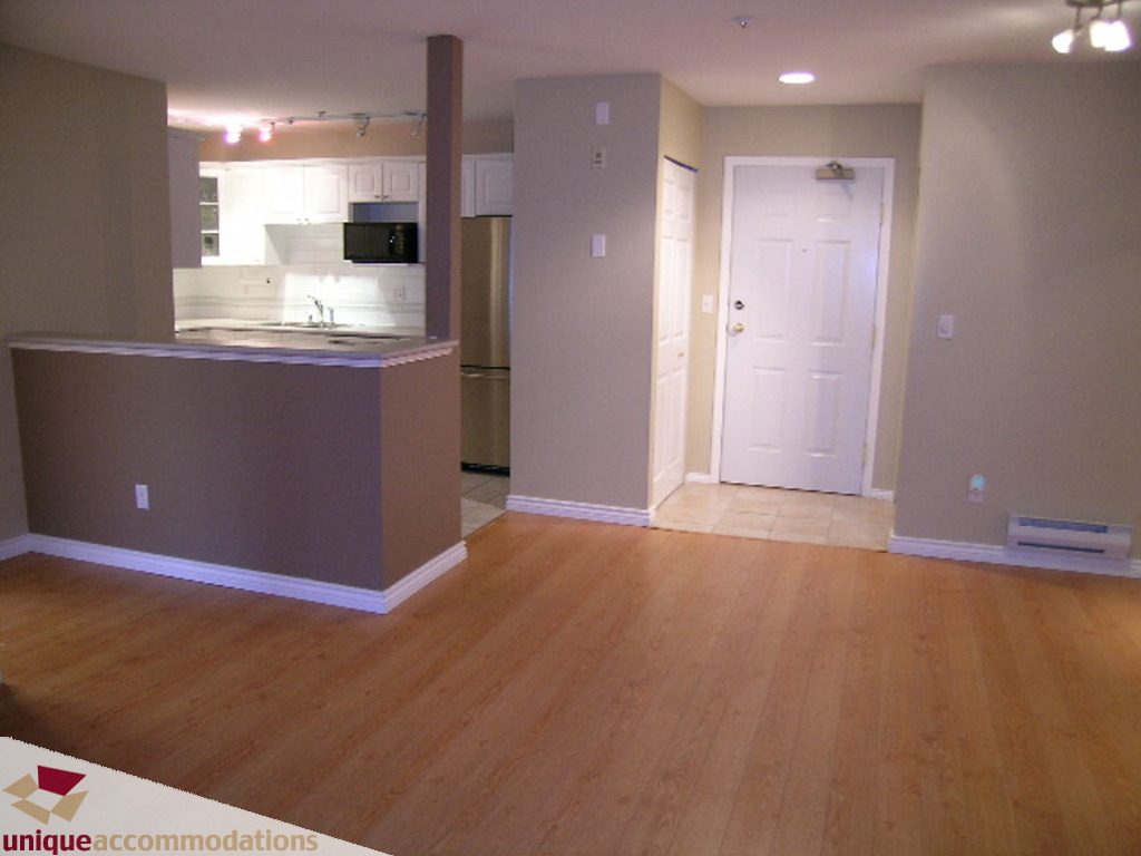 Open-concept living area with hardwood floors and a kitchen in the background. - 208 - 128 West 8th Street, North Vancouver