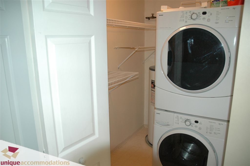 Bright laundry room with stacked washer and dryer, white shelving, and light-colored walls. - 208 - 128 West 8th Street, North Vancouver