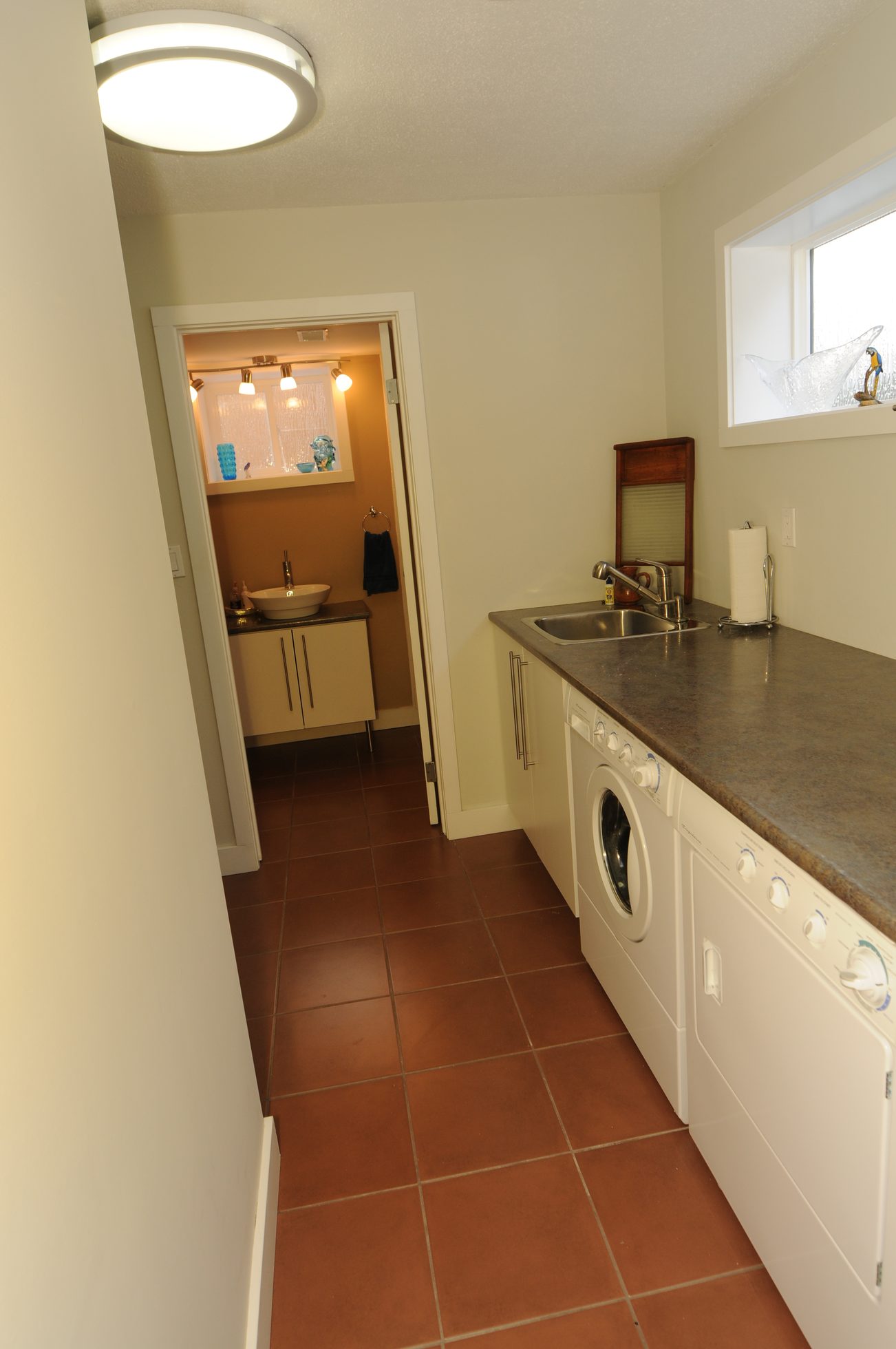 Bright laundry room with dark countertops and tiled floor, adjacent to a small bathroom with a sink and cabinet.