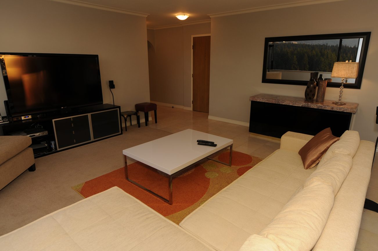 Furnished living room with a large TV, modern coffee table, and a view of the forest through a large window.