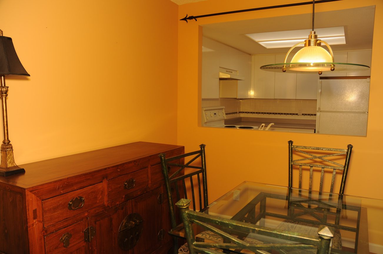 Bright dining area with wooden furniture and kitchen in the background, featuring a pendant light and yellow walls.