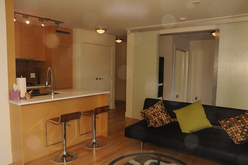 Furnished living room with black sofa, wooden bar stools, and modern kitchen counter. - 1405- 565 Smithe Street, Vancouver Downtown