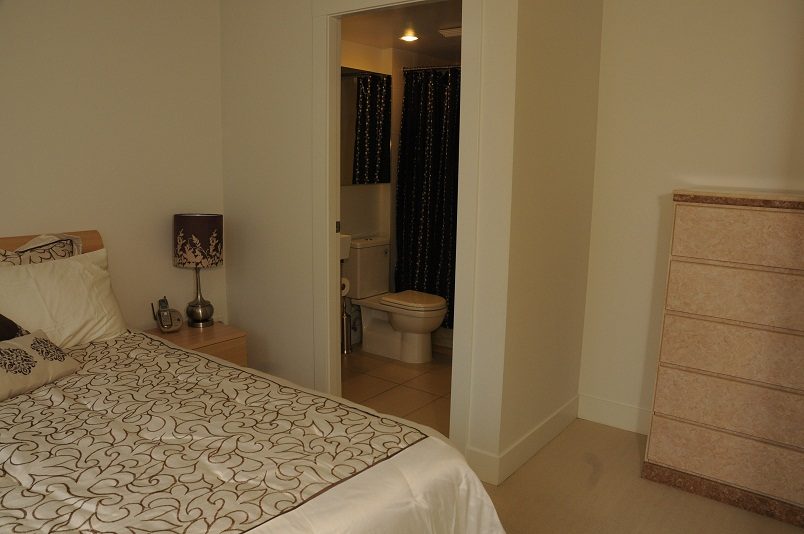 Bedroom with patterned bedding and nightstand with lamp, leading to a bathroom with dark curtains. - 1405- 565 Smithe Street, Vancouver Downtown