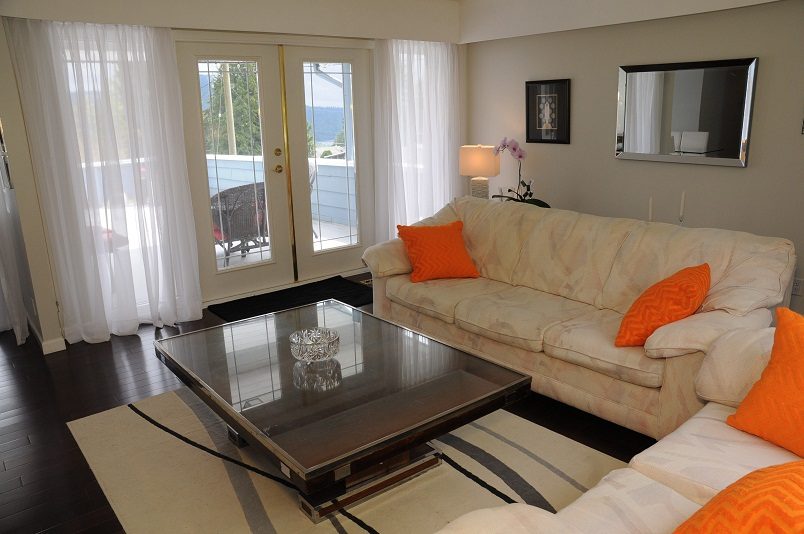 Furnished living room with white couch, orange pillows, glass coffee table, hardwood floors, and sliding glass doors...
