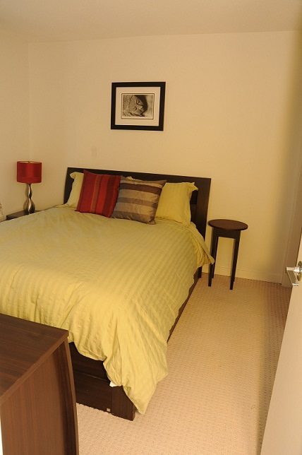 Furnished bedroom with yellow bedding and wooden headboard, small side table, and framed artwork on the wall. - 705 - 538 Smithe, Vancouver Downtown