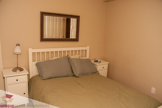 Furnished bedroom with neatly made bed, nightstand, and mirror. - 3035 Newmarket Drive, North Vancouver