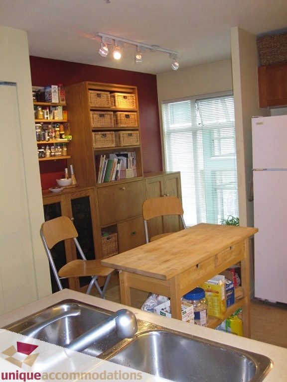 Bright kitchen with wooden table, chairs, and storage shelves, featuring a stainless steel sink and natural light from a...
