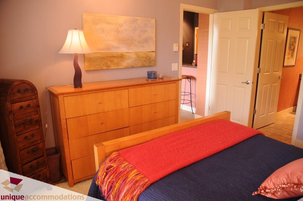 Furnished bedroom with wooden furniture, red accents, and a cozy ambiance. - 103-580 Raven Woods Drive, North Vancouver