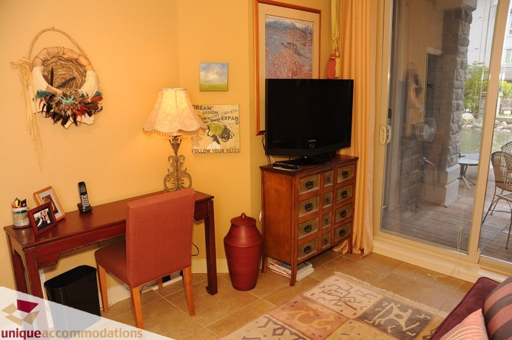Furnished living room with wooden desk, lamp, and sliding glass door to patio. - 103-580 Raven Woods Drive, North Vancouver