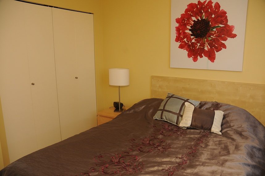 Furnished bedroom with a wooden headboard, brown bedding, and a vibrant red floral wall art. - 1501 -1077 Marinaside Crescent, Vancouver Downtown