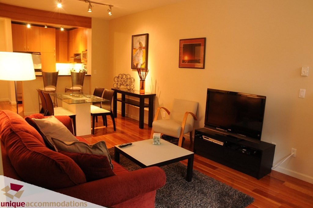 Furnished living room with hardwood floors, red couch, and wall-mounted TV. - 212- 580 Raven Woods Drive, North Vancouver