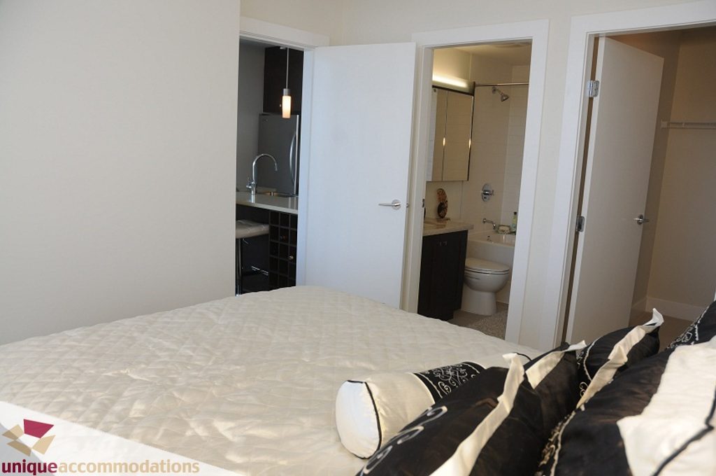 Furnished bedroom with white bedding and black accents, leading to a modern kitchenette and bathroom. - 3401 - 888 Homer Street, Vancouver Downtown