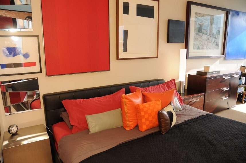Furnished 2-bedroom condo with vibrant red and orange accent pillows on the bed, modern artwork on the walls, and a sleek...
