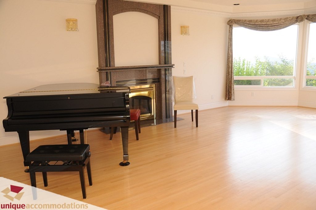 Bright living room with hardwood floors, piano, and fireplace, overlooking greenery. - 4139 Citadel Court, North Vancouver