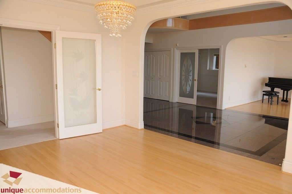 Bright living room with hardwood floors and a chandelier, leading to a dining area with a piano. - 4139 Citadel Court, North Vancouver