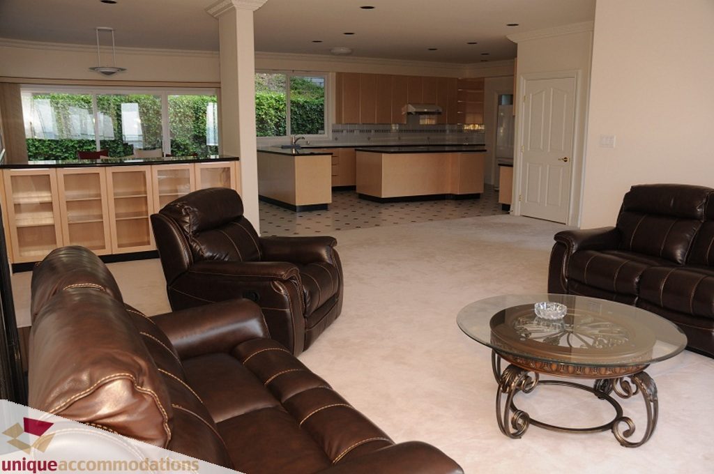 Spacious living room with leather furniture, glass coffee table, and kitchen in background. - 4139 Citadel Court, North Vancouver