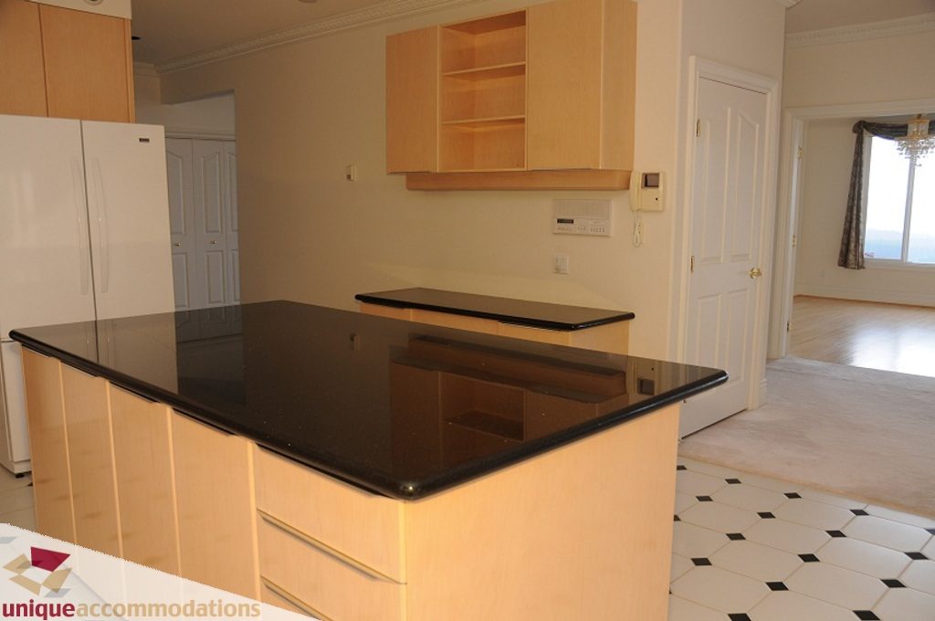 Bright kitchen with black countertops and light wood cabinets, leading to a spacious living area with a city view.