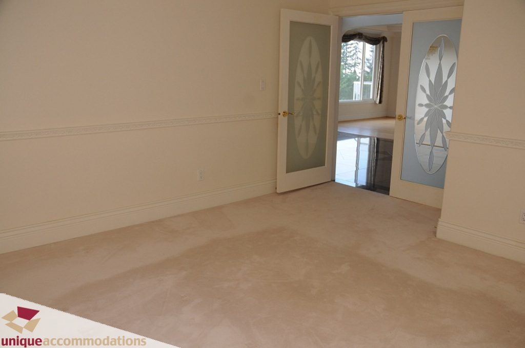 Empty living room with beige walls and carpet, leading to a door with decorative glass panels. - 4139 Citadel Court, North Vancouver