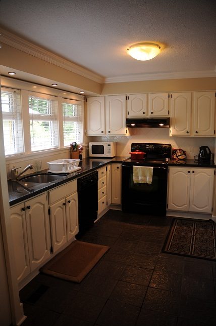 Bright kitchen with white cabinets, black appliances, and a window with blinds. - 6411 Nelson Avenue, West Vancouver