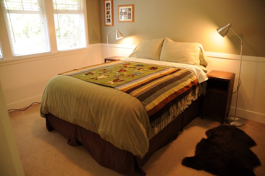 Bright bedroom with soft lighting and a cozy quilt, featuring a wooden nightstand and a window with blinds. - 6411 Nelson Avenue, West Vancouver