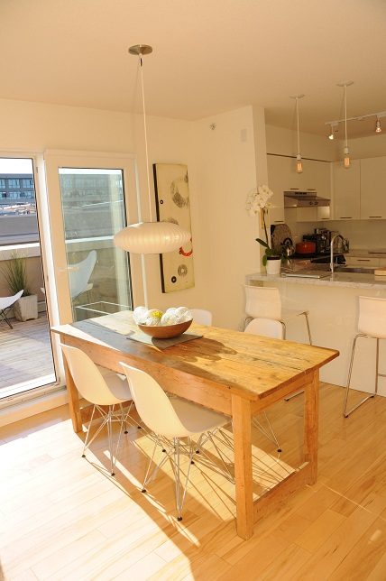 Bright dining area with wooden table, white chairs, and pendant lights, leading to a kitchen with white countertops and...