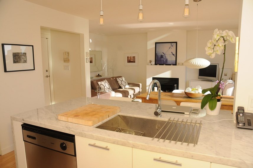 Bright kitchen with marble countertops and a modern sink, leading to a cozy living area with a fireplace and sofa.