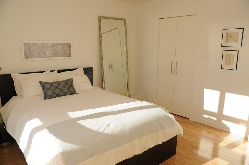 Bright bedroom with white walls, wooden floors, and a neatly made bed with white linens and patterned pillows.