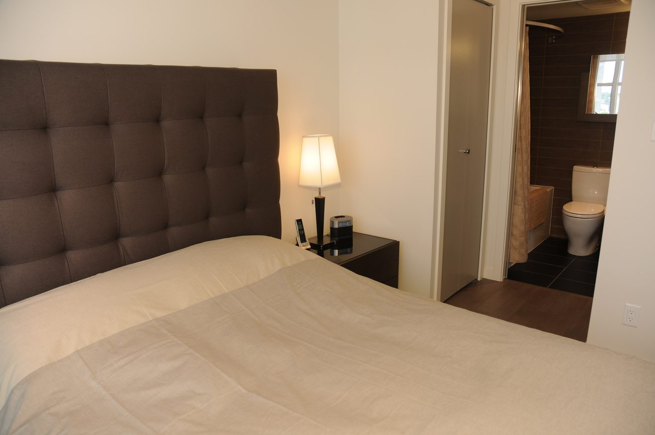 Furnished bedroom with tufted headboard and bedside lamp, leading to bathroom with toilet and shower. - 801 - 161 West Georgia, Vancouver Downtown