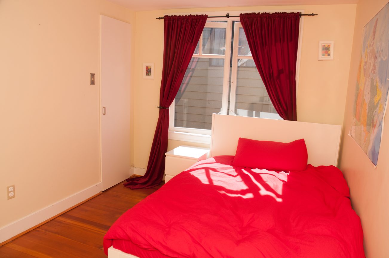 Bright bedroom with hardwood floors and red curtains, featuring a neatly made bed with red bedding. - 3249 West 36th Ave, Vancouver West Side