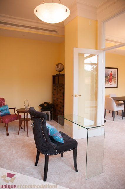 Bright living room with glass coffee table and plush armchair, yellow walls, and open door leading to dining area.