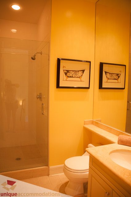 Bathroom with yellow walls, framed bathtub art, and double sink vanity. - 103-2288 Bellevue Ave, West Vancouver