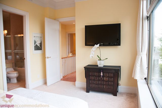 Bright living room with a mounted TV, open door leading to a bathroom, and a cozy corner with a bed and a small plant.