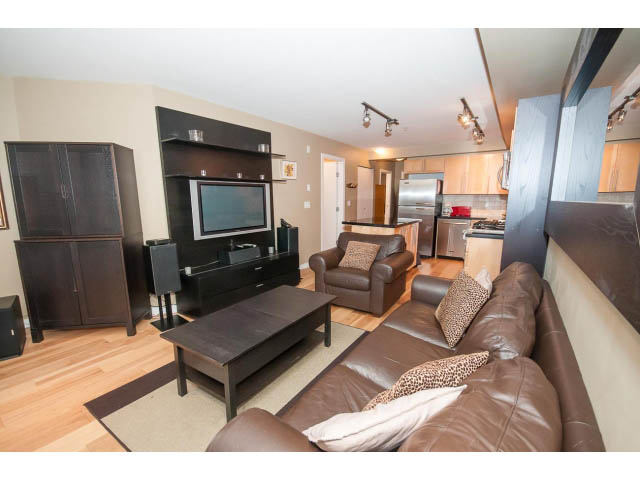 Furnished living room with hardwood floors, leather couch, and kitchen in background. - 204 - 122 East 3rd Street, North Vancouver