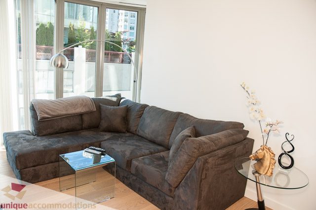 Furnished living room with large sectional sofa, glass coffee table, and city view through sliding doors. - 604-1000 Beach Avenue, Vancouver Downtown