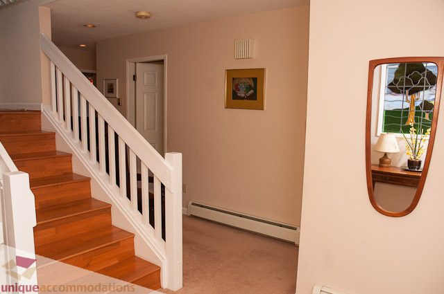 Bright hallway with wooden floors and white railings, leading to a door and a mirror reflecting the room. - 4559 W. 3rd Ave, Vancouver West Side