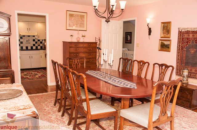 Furnished dining room with wooden chairs and a chandelier, leading to a kitchen with white cabinets and checkered backsplash.