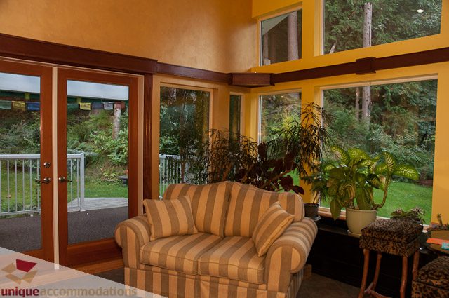Bright living room with large windows, lush greenery outside, and striped furniture. - 1650 Riverside Drive, North Vancouver
