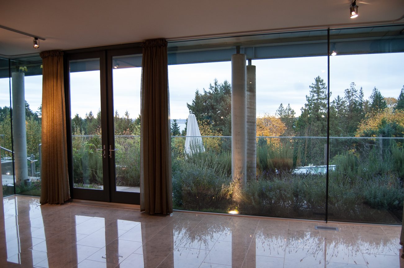 Bright living room with large glass windows, reflecting the lush greenery and a distant cityscape. - 4454 Regency Place, West Vancouver