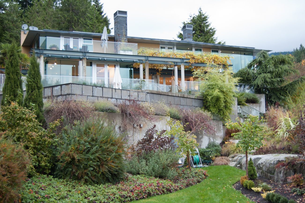 Spacious living room with glass walls and outdoor patio, surrounded by lush greenery. - 4454 Regency Place, West Vancouver