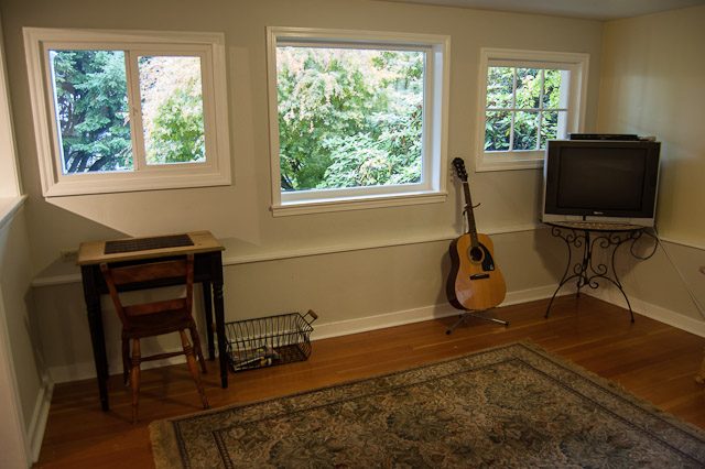 Bright living room with hardwood floors, a guitar, and a TV, featuring a cozy corner desk and large windows. - 139 Osbourne Rd. East, North Vancouver