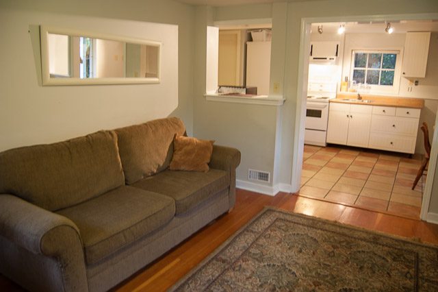 Furnished living room with brown couch, hardwood floors, and kitchen in the background. - 139 Osbourne Rd. East, North Vancouver