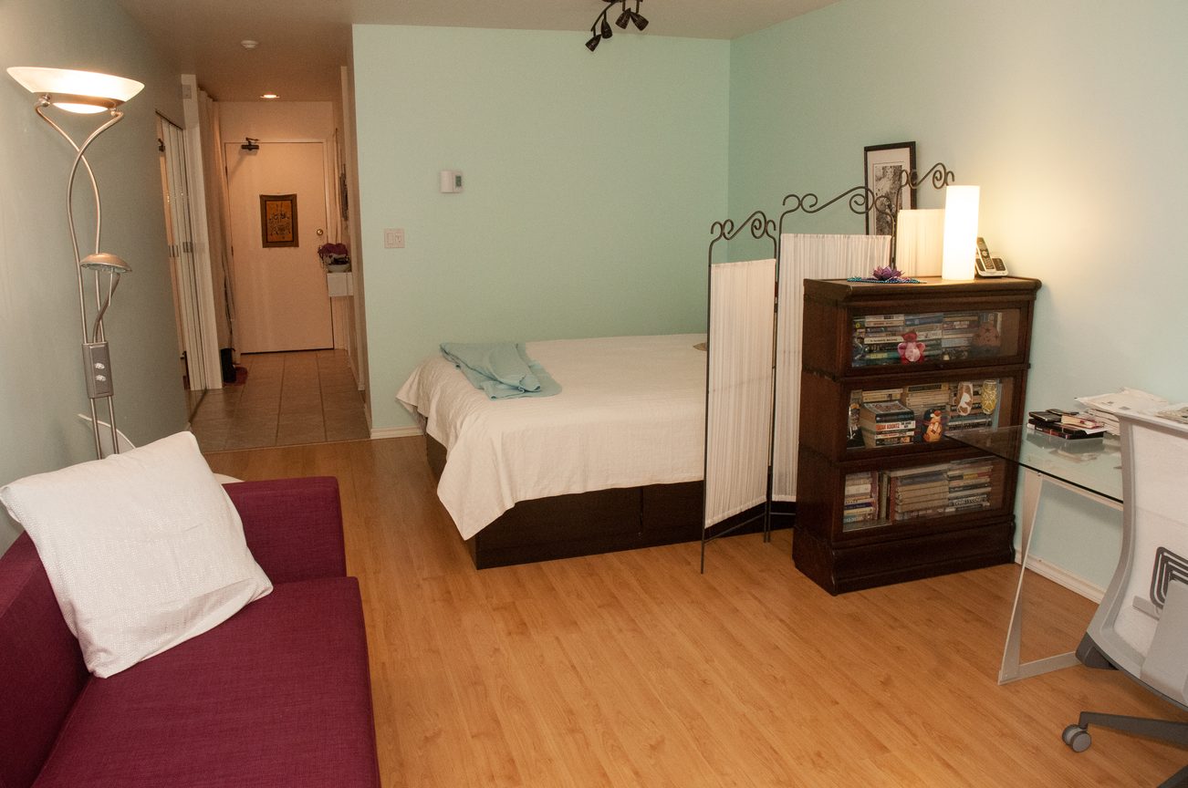 Furnished living room with hardwood floors, a red couch, and a small bedroom with a white bed and a bookshelf.