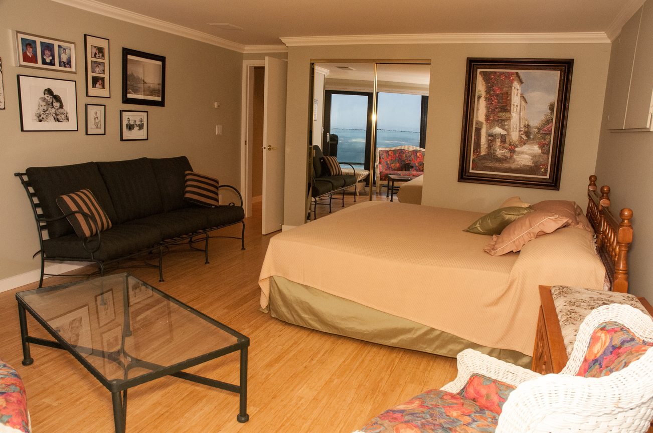 Spacious living room with hardwood floors, a large bed, and a sliding glass door leading to an outdoor patio with a city...