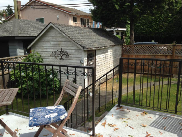 Furnished patio with metal railings and wooden chairs, leading to a shared house with a grassy yard and trees in the...